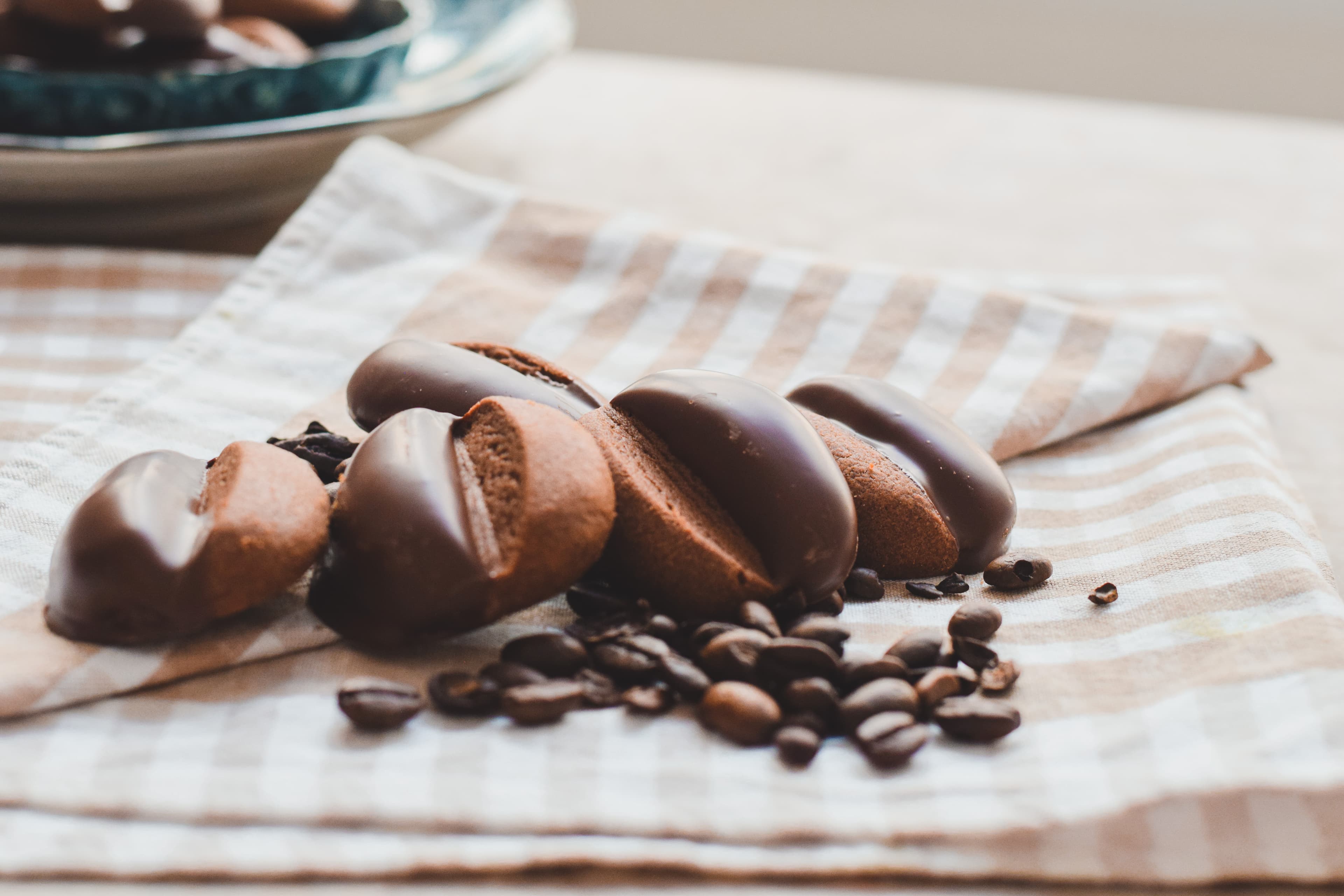 Chocolate-dipped Reena Bakes coffee cookies beside coffee beans on a striped cloth